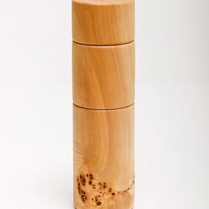 Pode incluir: Um recipiente cilíndrico de madeira clara, composto por três secções empilhadas, com tampa. A madeira apresenta um veio natural e um padrão decorativo de madeira de raiz na secção inferior. Altura aproximada: 15 cm.