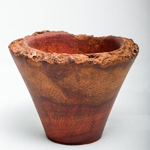 Può includere: Una ciotola decorativa conica fatta a mano con un bordo ruvido e strutturato. La ciotola presenta un gradiente di tonalità marrone-rossastro, che passa da una tonalità più chiara nella parte superiore a un tono più scuro alla base. L'interno ha una finitura liscia e rosso scuro.