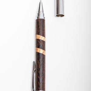 Könnte beinhalten: Ein Holzstift mit dunkelbraunem Finish und zwei hellbraunen Bändern. Der Stift hat einen silbernen Clip und eine Spitze, und die Kappe ist abgenommen und neben dem Stift platziert. Der Stift ist etwa 15 cm lang.