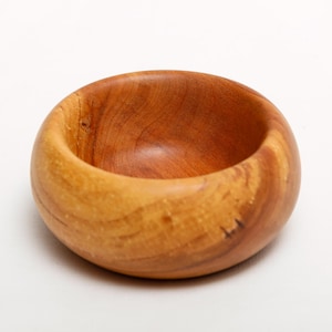 Könnte beinhalten: Eine kleine, runde Holzschale in einer natürlichen, warmen Farbe. Die Schale hat eine glatte, polierte Innenseite und eine etwas rauere Außenseite, die die Holzmaserung zeigt. Diese dekorative Schale ist ideal für kleine Gegenstände.
