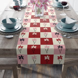 May include: A floral table runner with a checkered pattern in red, pink, and cream. Each square features a stylized flower in various colors. The runner is set on a wooden table with place settings and a vase of pink lilies.