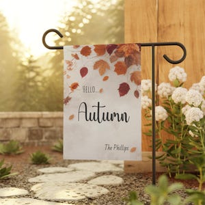 May include: A rectangular garden flag hanging on a black metal stand. The flag features a white background with a border of autumn leaves in shades of orange, red, and brown. The text reads "Hello... Autumn" in black script, with "The Phillips" below.