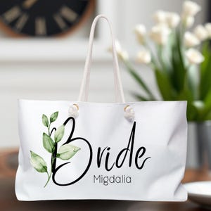 May include: White tote bag with a green leafy design and the word "Bride" in black cursive lettering. The name "Migdalia" is printed below the word "Bride".