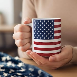 May include: A white ceramic mug featuring a printed design of the United States flag, displaying a blue field with white stars in the upper left corner and alternating horizontal red and white stripes covering the rest of the cylindrical surface.