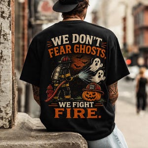 Peut inclure: Un t-shirt noir avec une impression graphique représentant un pompier pulvérisant de l'eau sur deux fantômes. Le texte indique WE DON'T FEAR GHOSTS, WE FIGHT FIRE. Une citrouille d'Halloween avec un casque de pompier est présente.
