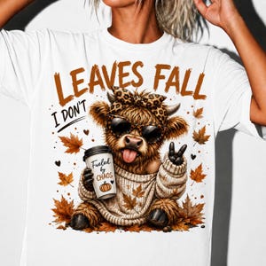 Peut inclure: Un t-shirt blanc avec un motif de vache Highland brune et poilue portant un bandeau à motif léopard et des lunettes de soleil. La vache porte un pull beige, tient un gobelet marqué Fueled by CHAOS et fait un signe de paix. Texte : LEAVES FALL I DON'T.