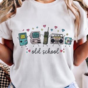 Peut inclure: Un t-shirt blanc avec une impression graphique de cinq appareils électroniques rétro anthropomorphes, dont une console de jeu portable, un ordinateur, un téléphone portable, un ghetto-blaster et un CD, avec le texte "old school" et des cœurs et étoiles colorés.