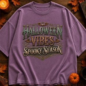 Peut inclure: Un t-shirt violet à manches courtes avec une impression graphique portant le texte HALLOWEEN VIBES SPOOKY SEASON dans une police stylisée. Le motif comprend des fioritures décoratives et de petites illustrations de chauves-souris.
