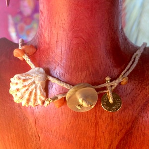 May include: A handcrafted necklace featuring a seashell, small orange beads, and a clear button-shaped pendant. The necklace also includes a small gold-colored disc pendant with text. The necklace is strung on a cream-colored cord.