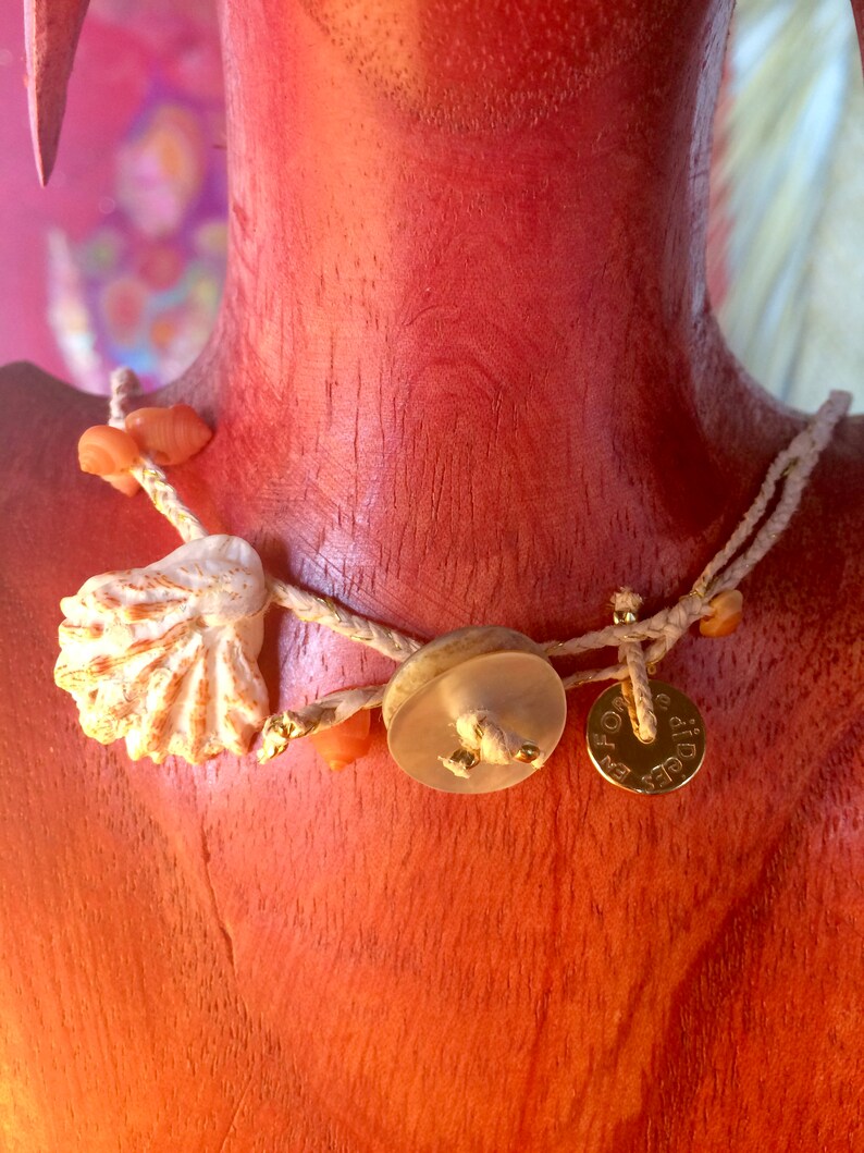 May include: A handcrafted necklace featuring a seashell, small orange beads, and a clear button-shaped pendant. The necklace also includes a small gold-colored disc pendant with text. The necklace is strung on a cream-colored cord.