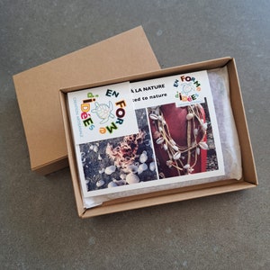 May include: A brown cardboard box with an open lid, revealing a printed card and a necklace. The card features images of seashells and text. The necklace is made of shells and twine. The box is on a gray surface.