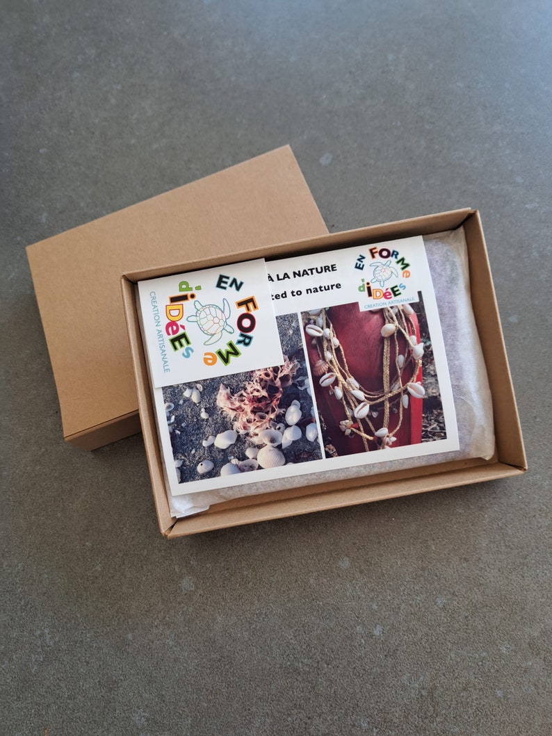 May include: A brown cardboard box with an open lid, revealing a printed card and a necklace. The card features images of seashells and text. The necklace is made of shells and twine. The box is on a gray surface.