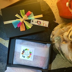 May include: Black gift boxes with colorful paper bows and tags. One box is open, revealing a wrapped item. The tags read "en forme d'id&eacute;es". The boxes are on a dark surface, with a decorative shell and a red box in the background.