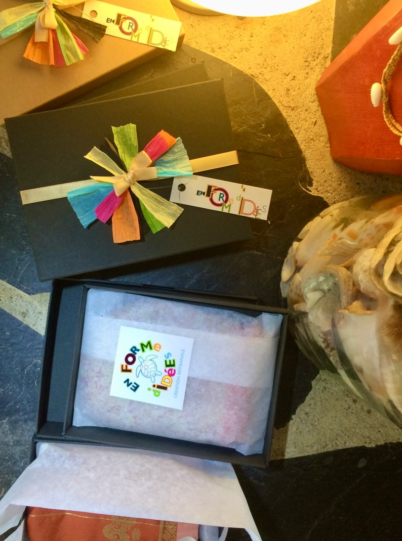 May include: Black gift boxes with colorful paper bows and tags. One box is open, revealing a wrapped item. The tags read "en forme d'id&eacute;es". The boxes are on a dark surface, with a decorative shell and a red box in the background.