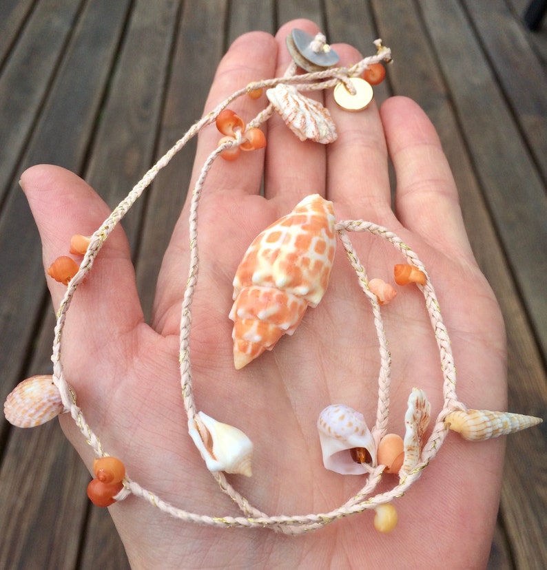 May include: A seashell necklace featuring a large, patterned orange and white shell pendant. The necklace includes various smaller shells, orange beads, and gold accents, strung on a cream-colored cord. The necklace is displayed on a wooden surface.