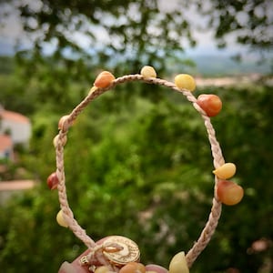 Peut inclure: Un bracelet tressé orné de perles marron clair et jaunes. Le bracelet est composé d'un cordon tressé beige et d'un fermoir doré. L'arrière-plan est une vue floue d'arbres et de feuillage verts.
