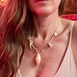 May include: A cream-colored necklace featuring various seashells and small beads strung on a white cord. The focal point is a large, textured shell pendant. The necklace is worn against a light-colored top.