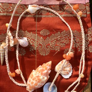 May include: A necklace made of braided rope and seashells. The necklace features a variety of seashells in shades of orange, white, and tan. The necklace is displayed on a red and gold patterned fabric.