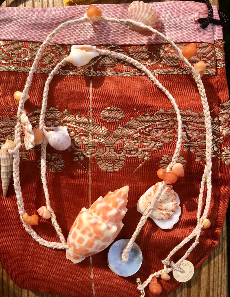 May include: A necklace made of braided rope and seashells. The necklace features a variety of seashells in shades of orange, white, and tan. The necklace is displayed on a red and gold patterned fabric.