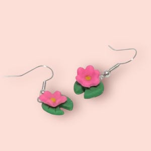 Peut inclure: Une paire de boucles d'oreilles faites à la main avec des fleurs roses, des centres jaunes et des feuilles de nénuphar vertes. Les boucles d'oreilles ont des crochets argentés. Sur un fond rose clair, ces boucles d'oreilles sont un accessoire charmant.