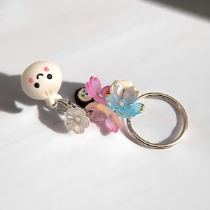 May include: A silver-toned metal keychain featuring a small, round, white dumpling charm with a smiling face and pink cheeks, accompanied by several translucent, multi-colored flower charms in shades of pink, blue, and clear, plus a small sushi charm.