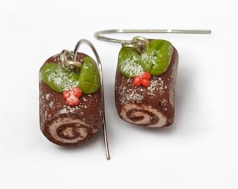May include: A pair of dangle earrings shaped like miniature chocolate yule log cakes. Each brown cake features a white spiral center, green holly leaves, and small red berries on top, dusted with white powder and attached to silver hooks.