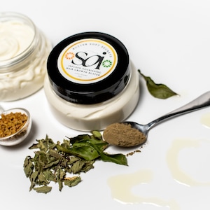 May include: Two jars of white hair growth butter with a black label that reads "Infused Nourishing Hair Growth Butter" and a green and yellow logo that says "Sai". The jars are surrounded by dried herbs, a spoon with brown powder, and a small measuring spoon with brown seeds.