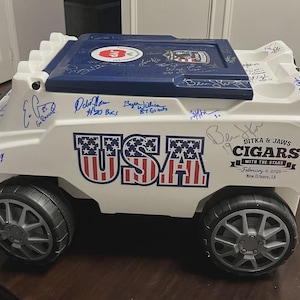 Puede incluir: Una nevera blanca y azul con ruedas negras, con la palabra "USA" en rojo, blanco y azul. La nevera tiene firmas y el texto "DITKA & JAWS CIGARS WITH THE STARS February 6, 2025 New Orleans, LA."