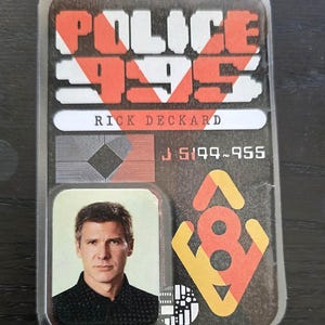 May include: A rectangular ID badge with the text "POLICE 995" in red and white. The badge includes a photo of a man, the name "RICK DECKARD", and the numbers "J 5199-955". The badge has a retro design.