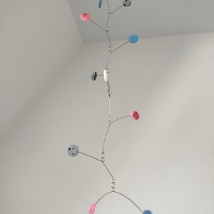 Könnte beinhalten: Eine moderne Mobile-Skulptur mit einem filigranen, ausgewogenen Design. Sie besteht aus einer Reihe von Drahtarmen, die farbige, kreisförmige Elemente in Rosa, Blau und gesprenkeltem Grau aufhängen. Das Mobile hängt an einem einzigen Punkt und erzeugt ein Gefühl von Bewegung und künstlerischem Flair.