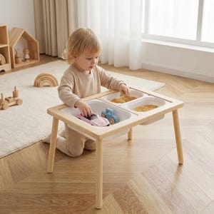 May include: A light wood sensory table with three white plastic bins. The table has four legs and a rectangular frame. The bins contain small toys and grains. A child is playing with the table and toys.