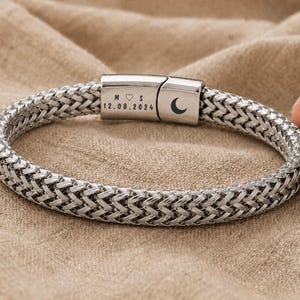 Puede incluir: Una pulsera de cadena trenzada en tono plateado con un cierre metálico rectangular. El cierre está grabado con las letras M y S, un símbolo de corazón, la fecha 12.08.2024 y un icono de media luna en el lateral.