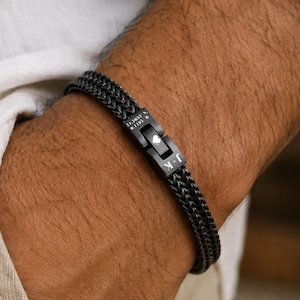 May include: A black double-strand wheat chain bracelet featuring a rectangular clasp. The clasp is engraved with the text 51.3833 N, 0.1195 W, a small heart symbol, and the letters J + K in a clean, white, sans-serif font style.