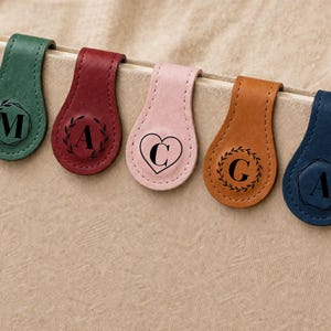 Pode incluir: Cinco marcadores de couro em verde, bordô, rosa, castanho claro e azul marinho, cada um com uma inicial e um elemento decorativo diferente. Os marcadores estão presos no topo de um livro.