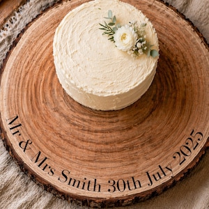 May include: A round, frosted cake decorated with white flowers and greenery sits on a rustic wooden serving board. The board is engraved with the text "Mr & Mrs Smith 30th July 2025". The cake is a light cream color.
