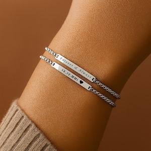 Può includere: Due braccialetti d'argento sono mostrati, ciascuno con una placca rettangolare e una catena a maglie. Il braccialetto superiore recita "Forever & Always", e quello inferiore "IX.IV.MMI" con un simbolo a cuore.