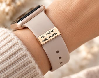 May include: A light beige silicone smartwatch band featuring a rectangular gold-toned metal charm engraved with the black text YOU CAN DO HARD THINGS. The band has a smooth finish with circular adjustment holes and a matching gold-toned watch case.