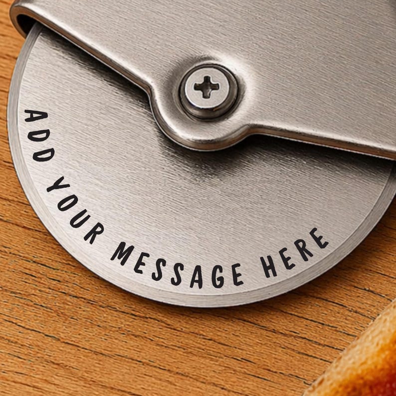 May include: Close-up of a stainless steel pizza cutter with the text "ADD YOUR MESSAGE HERE" etched on the blade. The cutter is on a wooden surface, with a portion of pizza visible.