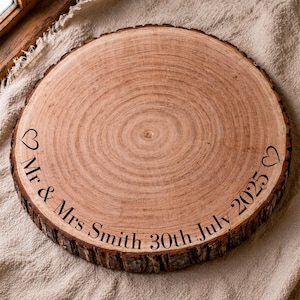 May include: A round, wooden tree slice with a natural bark edge. The surface is engraved with the words "Mr & Mrs Smith 30th July 2025" and two heart symbols. The wood has a light brown color.