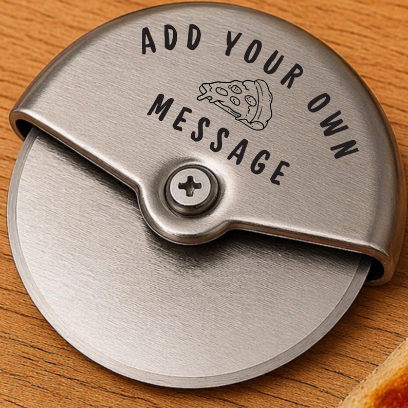 May include: A stainless steel pizza cutter with a curved handle and a circular blade. The handle features the text "ADD YOUR OWN MESSAGE" with a pizza slice graphic. The cutter is on a wooden surface.