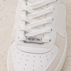 May include: Close-up of a white sneaker with white laces and a silver tag that reads "YOU GOT THIS." The shoe has a perforated design on the toe and a suede-like material on the side.