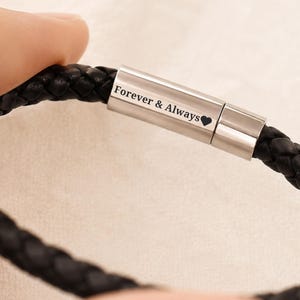 May include: A black braided leather bracelet featuring a cylindrical silver-toned metal clasp. The clasp is engraved with the text Forever & Always followed by a small heart symbol. The bracelet has a smooth, polished finish on the metal component.