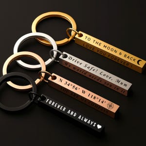 Può includere: Una collezione di portachiavi rettangolari in oro, argento, oro rosa e nero. Ogni portachiavi ha un anello e testo inciso. Il testo include frasi come "To the moon & back" e "Forever and always".