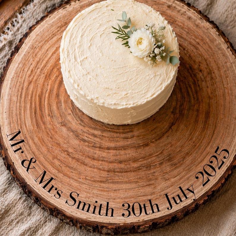 May include: A round, frosted cake decorated with white flowers and greenery, placed on a rustic wooden serving board. The board is engraved with the text "Mr & Mrs Smith 30th July 2025". The cake has a textured frosting.