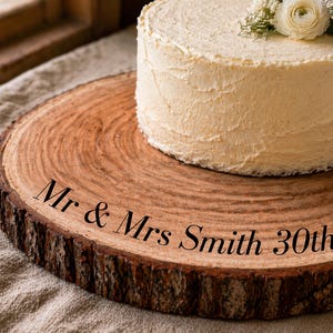 May include: A round, frosted cake with floral decorations sits atop a rustic wooden cake stand. The cake stand is a cross-section of a tree trunk, with the words "Mr & Mrs Smith 30th Ju" inscribed on it. The cake has a creamy white frosting.