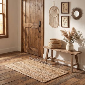 May include: A rectangular woven jute rug with a natural tan texture lies on a wooden floor. The text PureloomRugs is visible on the rug. A wooden bench sits nearby, holding two stacked round woven baskets and a ceramic vase with dried foliage.