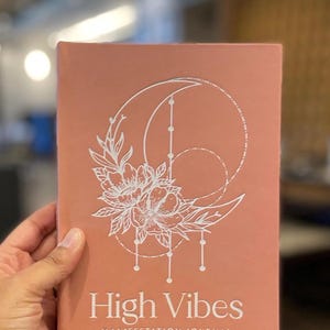 May include: A dusty rose-colored journal with a white line-art graphic featuring a crescent moon, circular geometric elements, and floral details. The text "High Vibes" is printed in a large serif font above "MANIFESTATION JOURNAL" in smaller caps.
