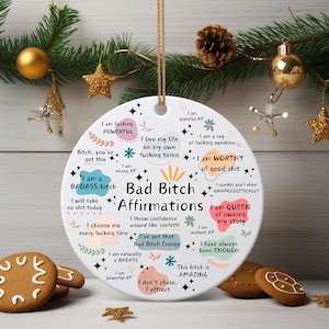Könnte beinhalten: Ein runder, weißer Keramikschmuck mit dem Text "Bad Bitch Affirmations" und motivierenden Sätzen in Schwarz und Rosa. Der Schmuck wird an einer Schnur aufgehängt. Festliche Weihnachtsdekorationen im Hintergrund.