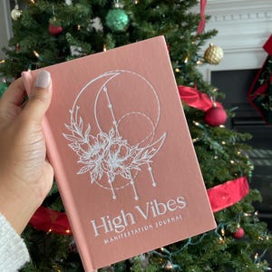 Pu&ograve; includere: Un diario con copertina rigida color rosa polvere con un'illustrazione a linee bianche di una luna crescente e fiori. Il testo "High Vibes" &egrave; stampato in grassetto bianco sopra il testo pi&ugrave; piccolo "MANIFESTATION JOURNAL" sulla copertina.