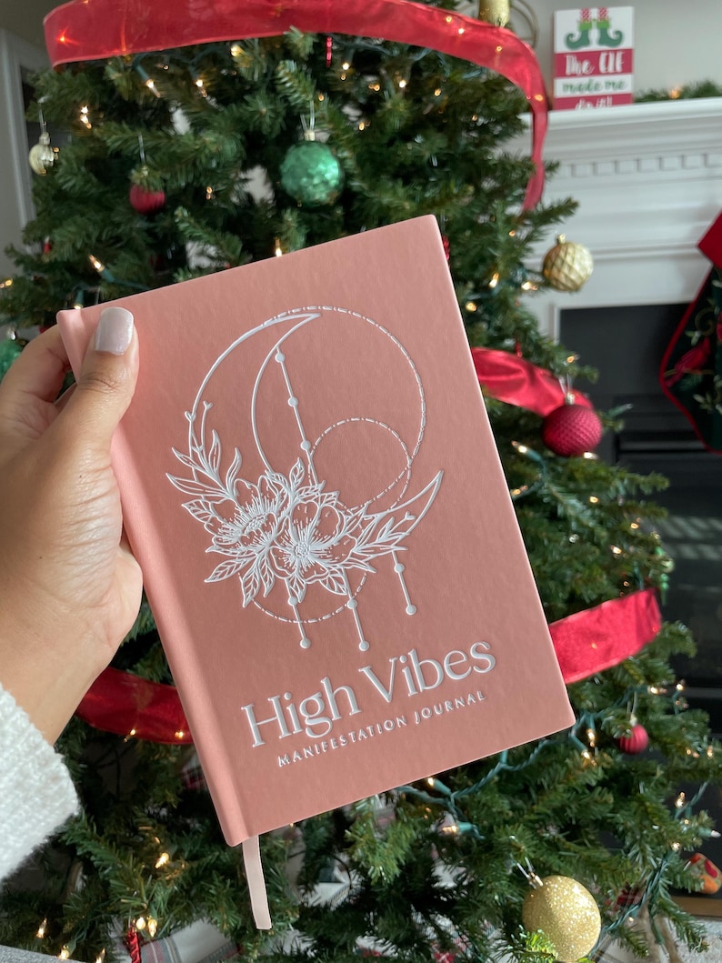 Pu&ograve; includere: Un diario con copertina rigida color rosa polvere con un'illustrazione a linee bianche di una luna crescente e fiori. Il testo "High Vibes" &egrave; stampato in grassetto bianco sopra il testo pi&ugrave; piccolo "MANIFESTATION JOURNAL" sulla copertina.
